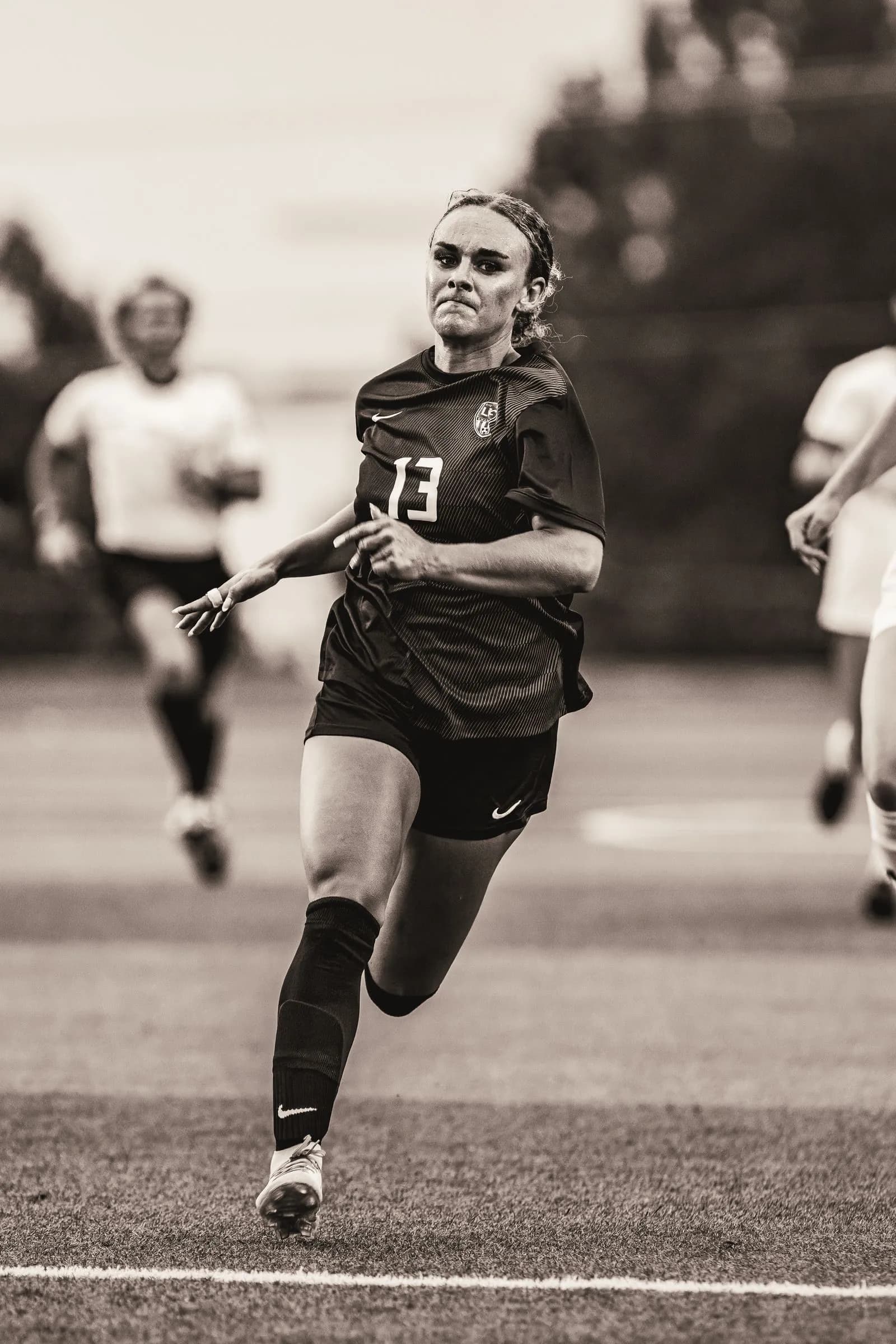 A female athlete in a dark jersey number 13 runs on an outdoor field marked with a white line under bright daylight.
