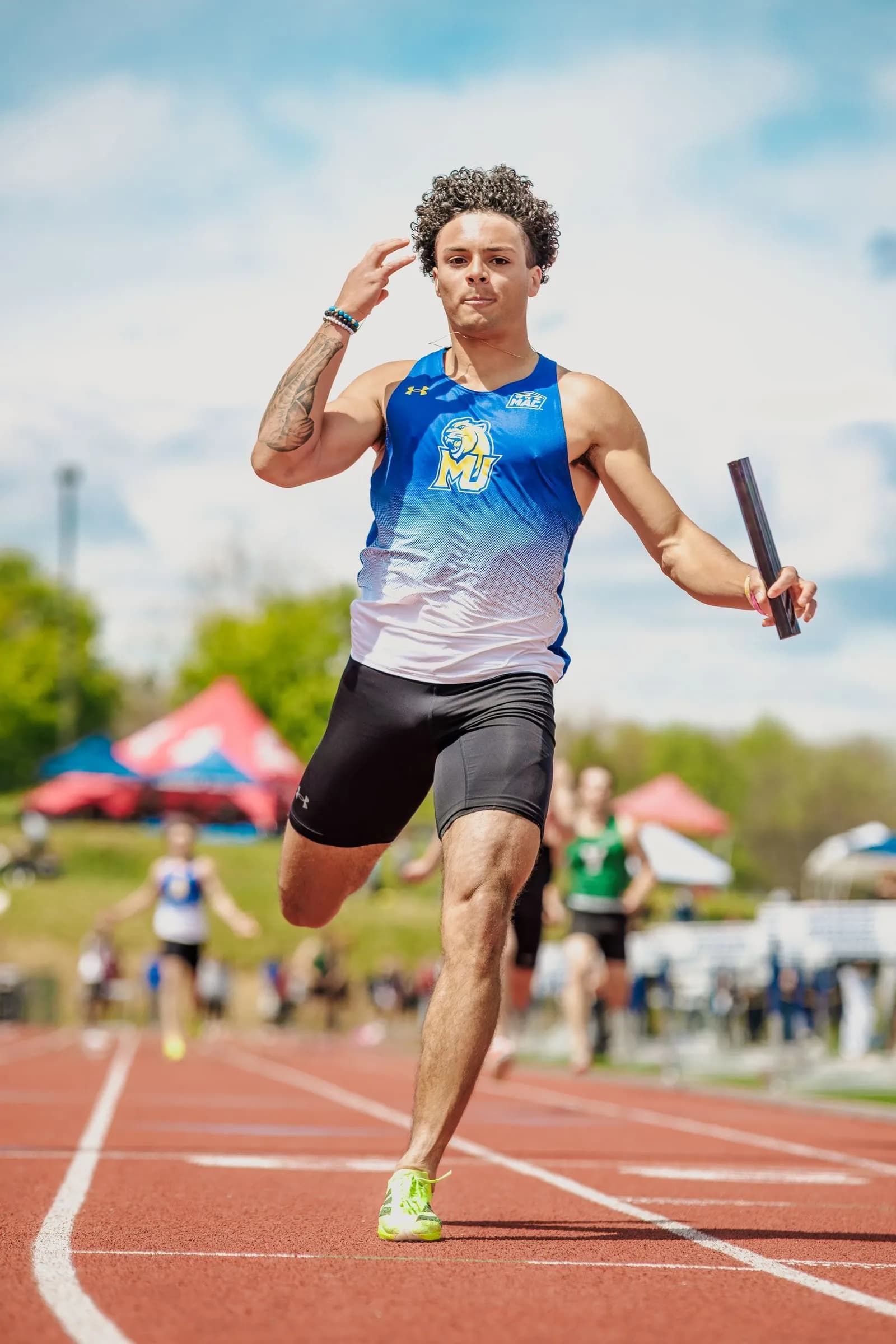 Male athlete runs on an outdoor track holding a relay baton, with other runners and spectators behind him.