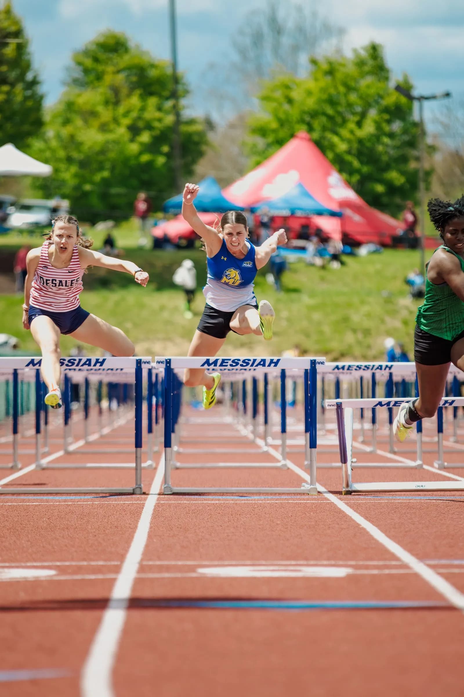 Three female athletes hurdle on an outdoor track beside tents and trees, one soaring mid‑air over a hurdle.