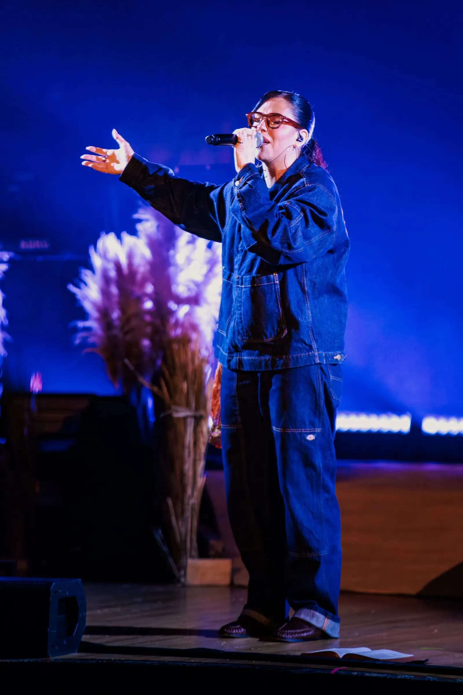 Lizzie Morgan, in a denim jacket and jeans, reaches out an arm while singing on a blue-lit stage with pampas grass decor