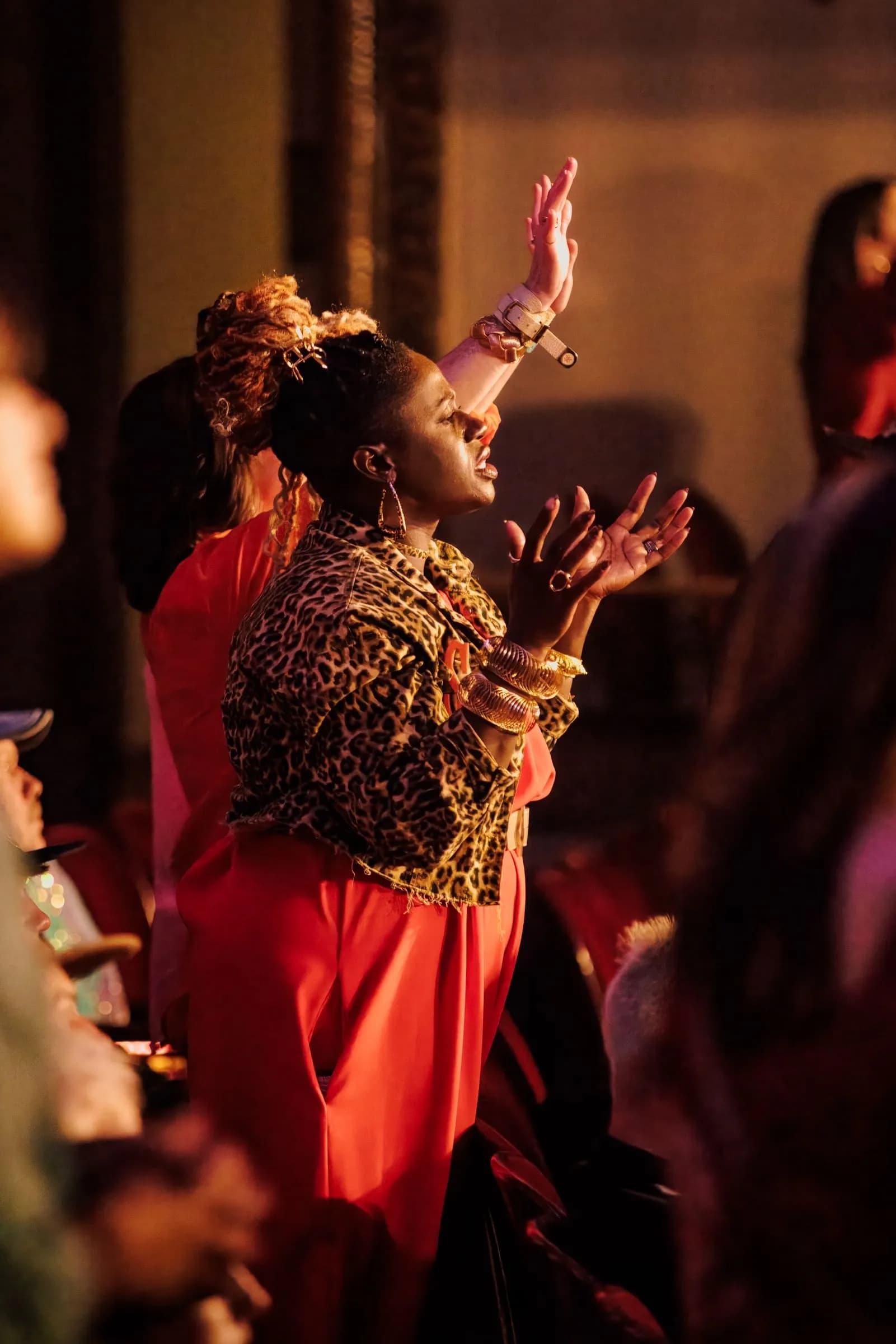A backing vocalist in a leopard-print jacket worships with hands raised in warm amber light