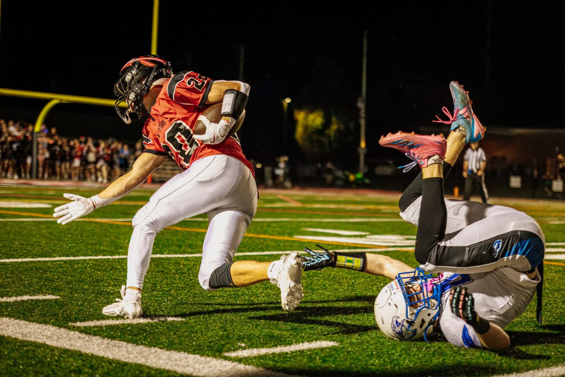 Two football players clash as one runs forward and the other lies on the field under bright stadium lights at night.