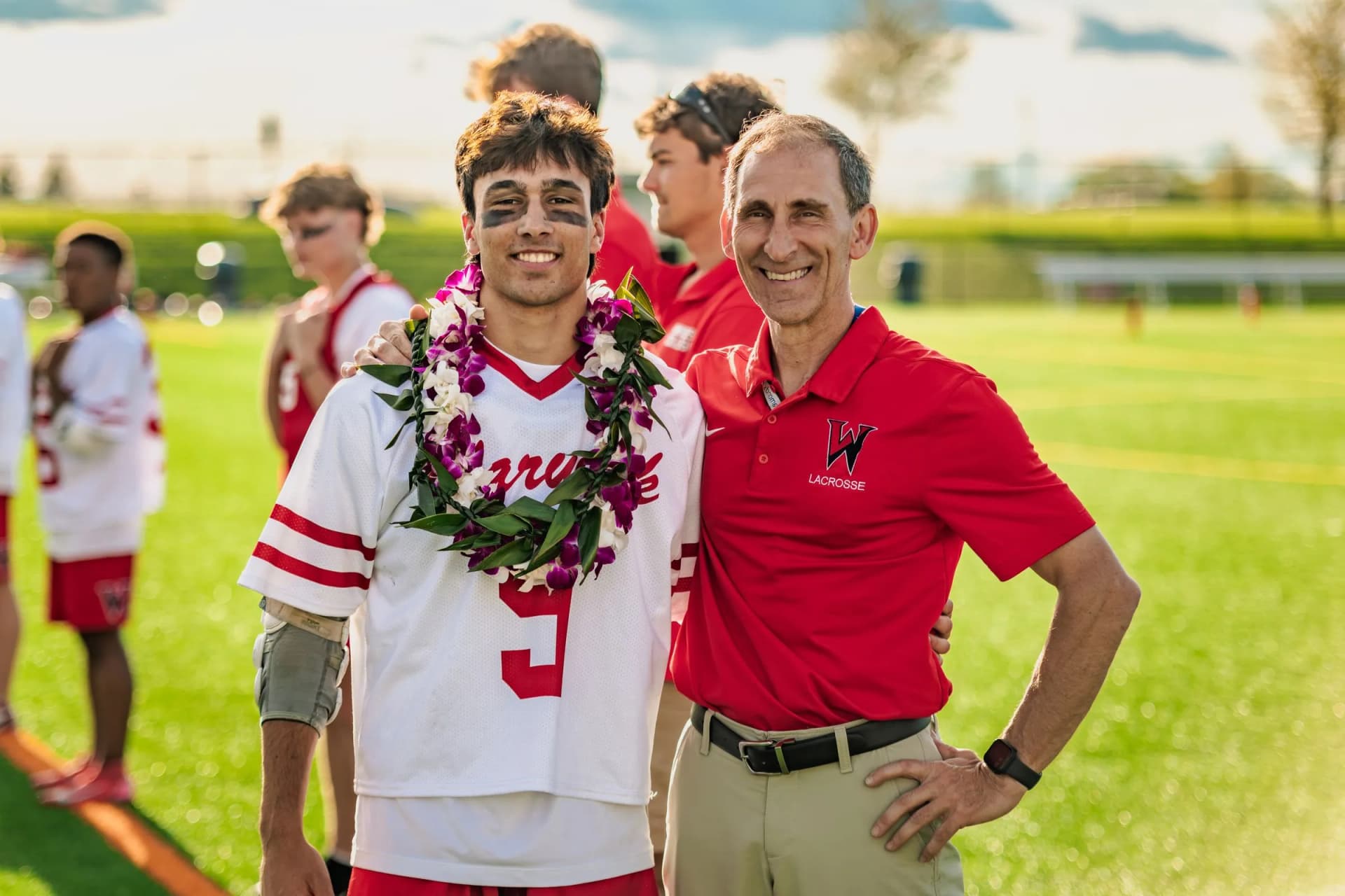 Two men stand side by side posing on a bright grassy field while other athletes watch from the background.