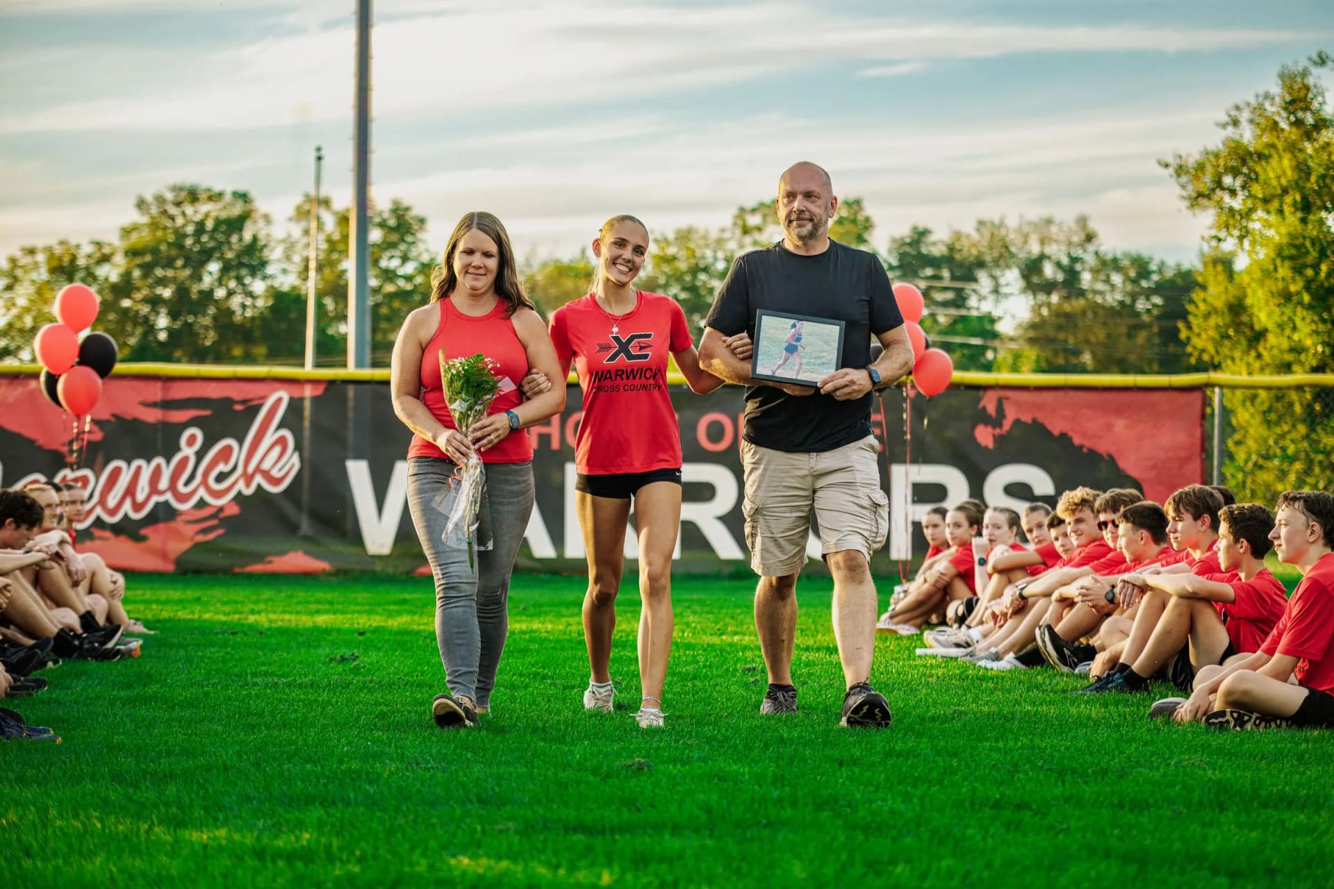 Three people walk forward across a grass field, a man carrying a framed picture and a woman holding a bouquet, outdoors.