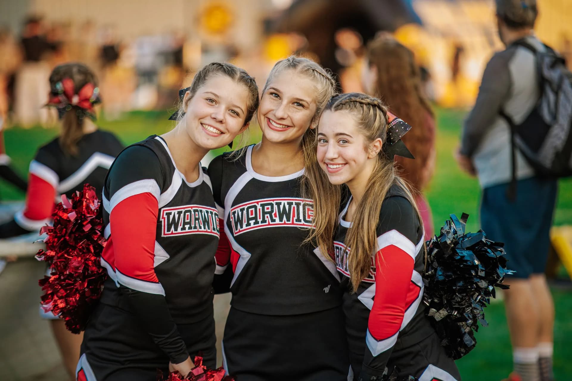 Three cheerleaders pose together on a green field in a stadium, with a blurred crowd of spectators behind them.