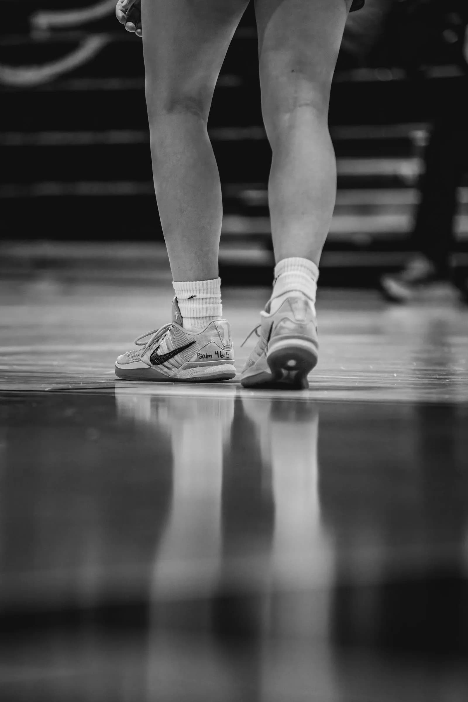 Legs and feet in athletic shoes stand on a highly reflective indoor floor, showing clear reflections of the legs.