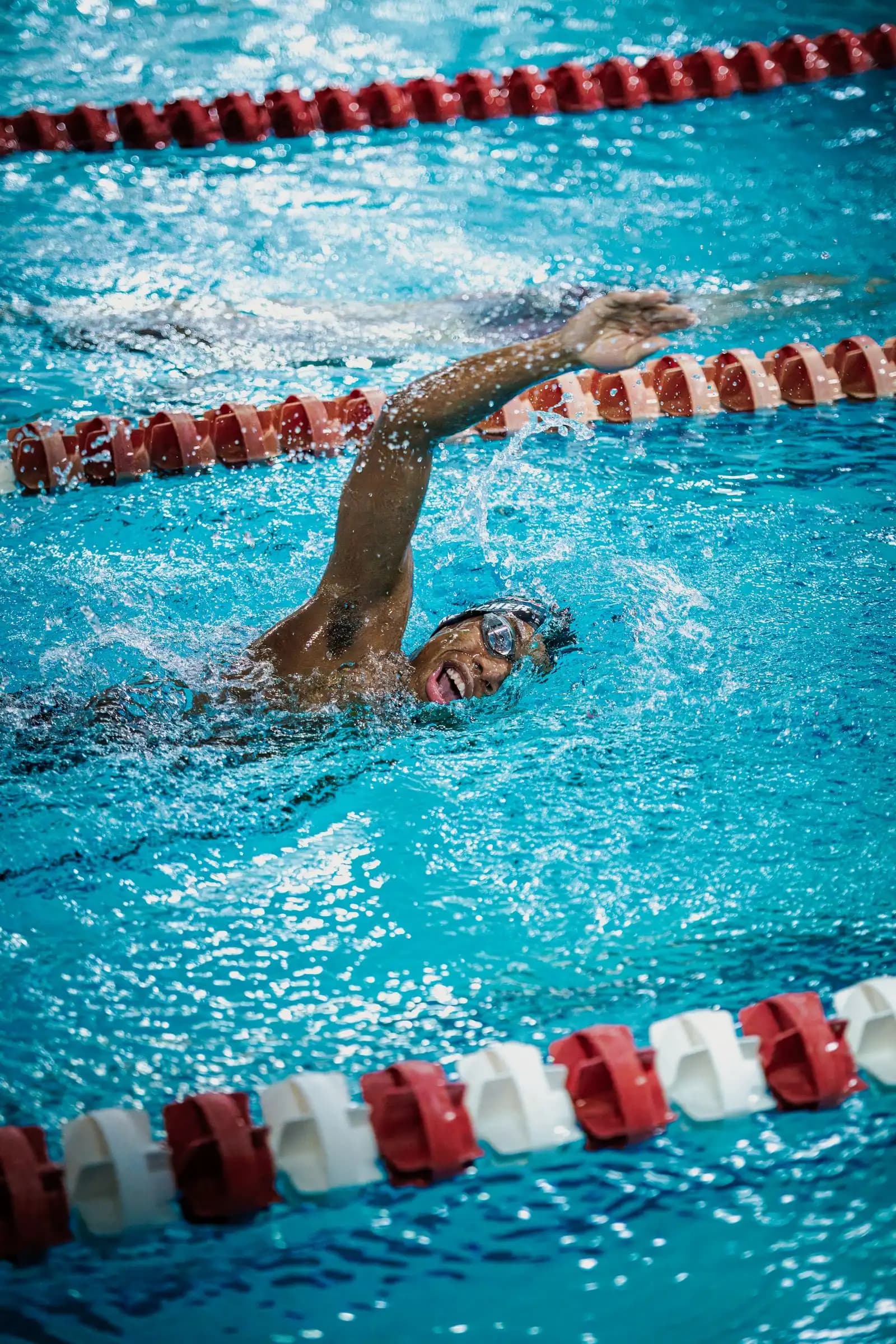 A swimmer in goggles strokes powerfully with an arm raised, mouth open, under bright indoor pool lighting.