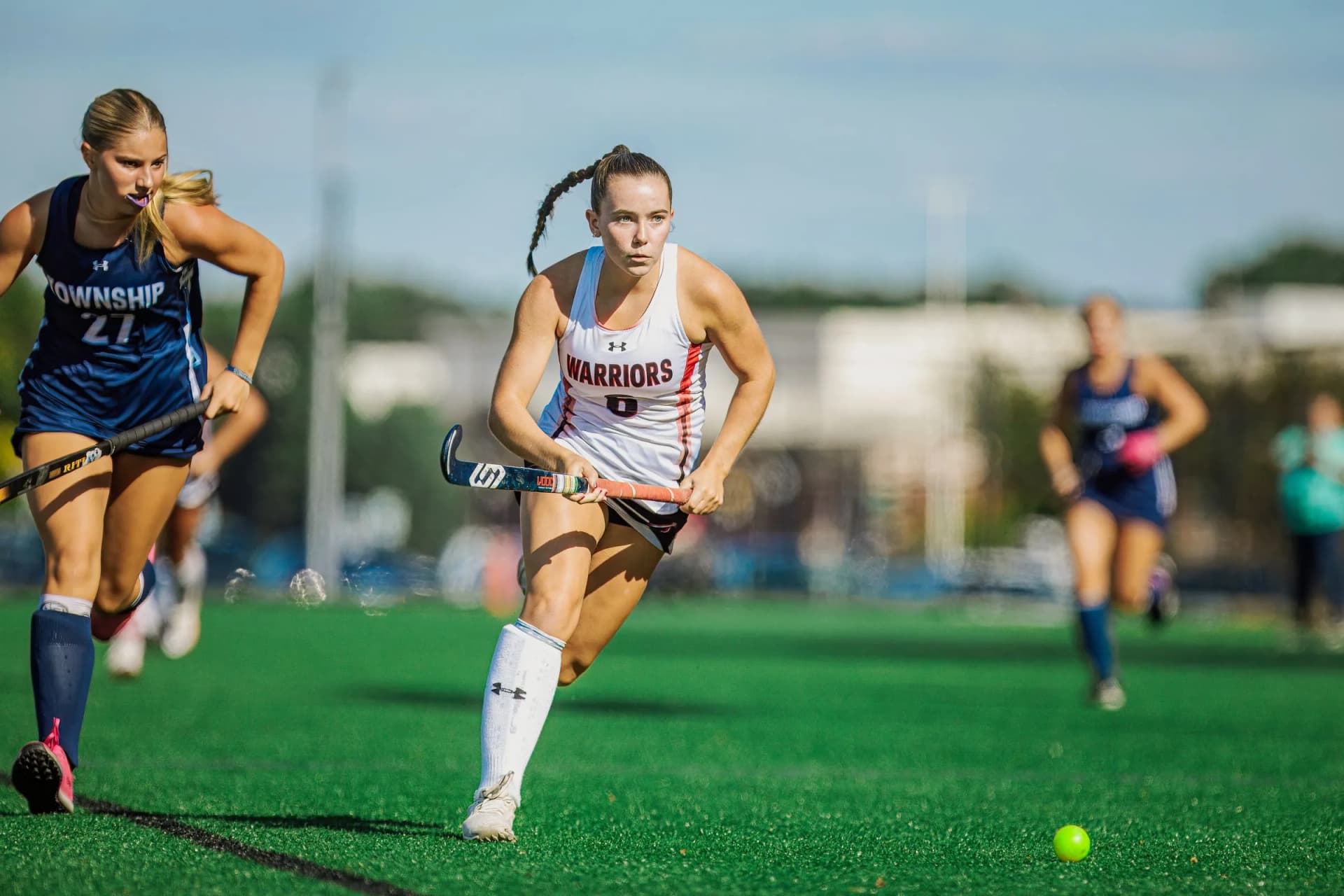 Female field hockey players sprint across a green turf field outdoors, one in a white jersey holding a stick near a ball.