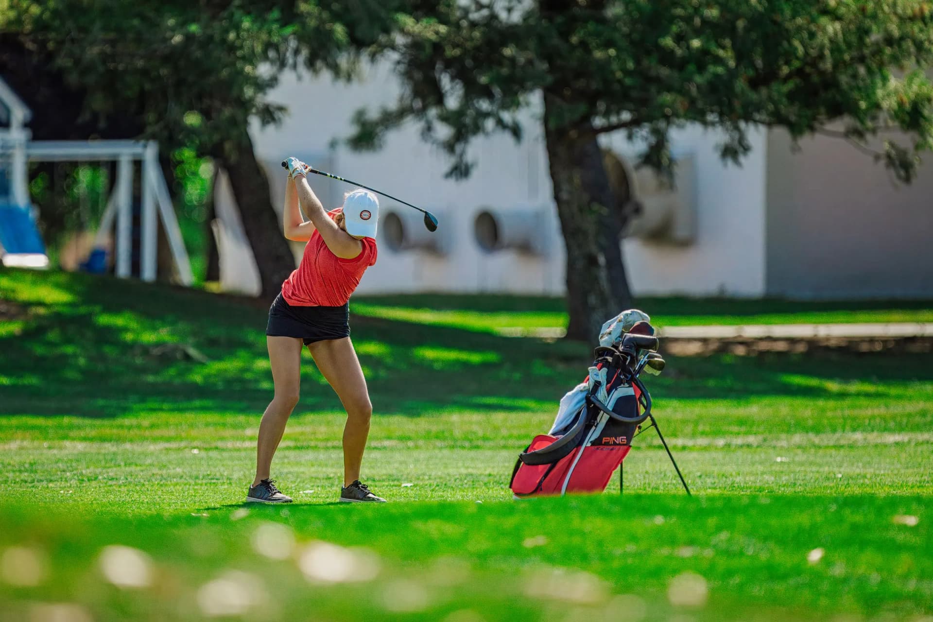 A person in a red shirt swings a golf club on an outdoor grass course beside a white building and trees.