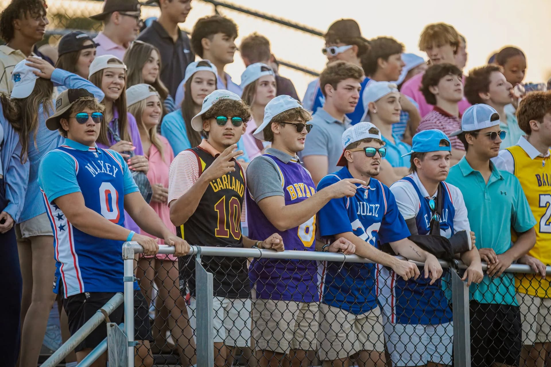 A dense crowd of young spectators stands behind a metal railing, all looking toward the right at an outdoor sports venue.