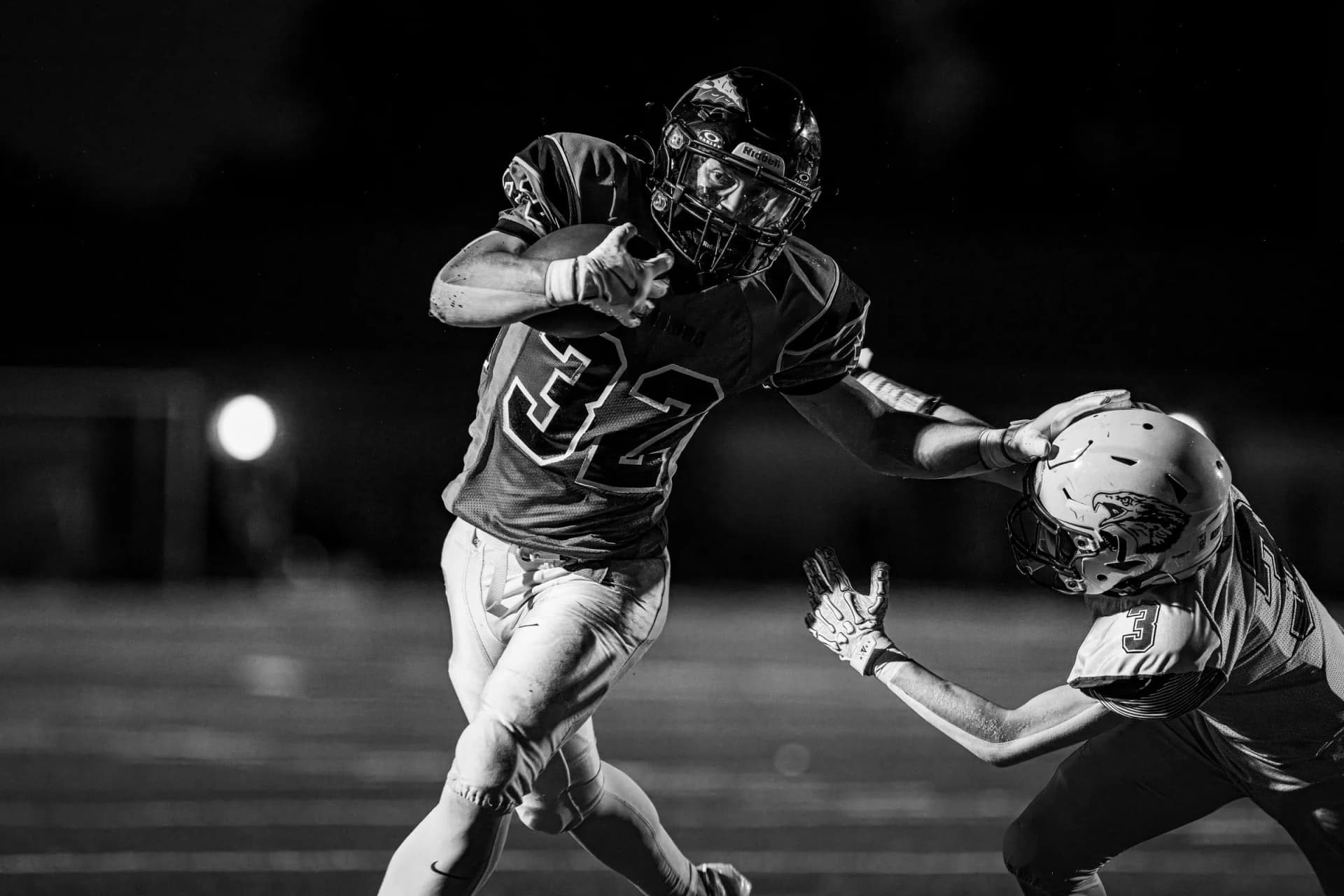 Two football players in full gear sprint on a night field, the ball carrier extending his arm toward a crouching defender.