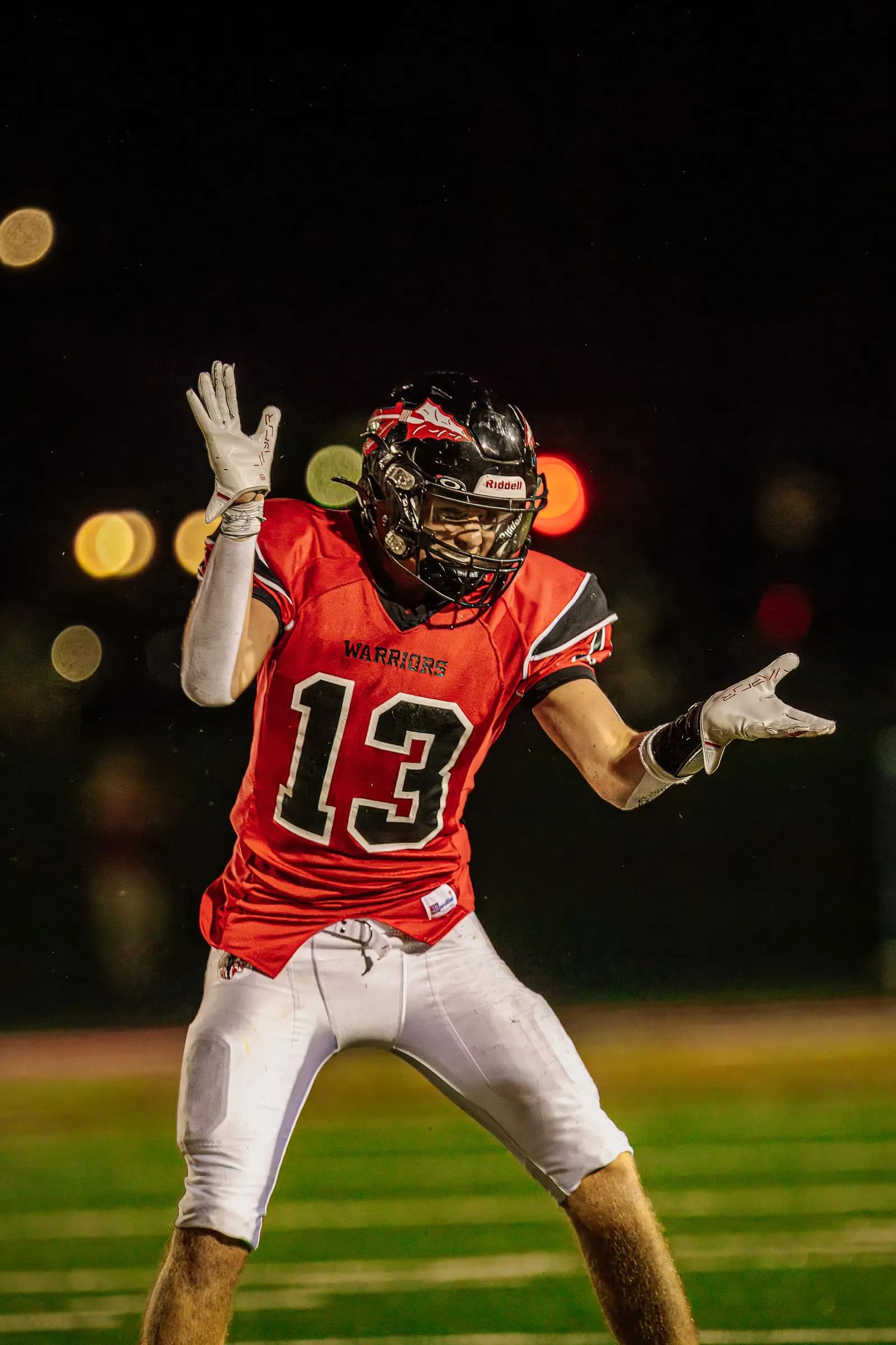A football player with arms outstretched stands on an outdoor night field as another player's arm rises in front.