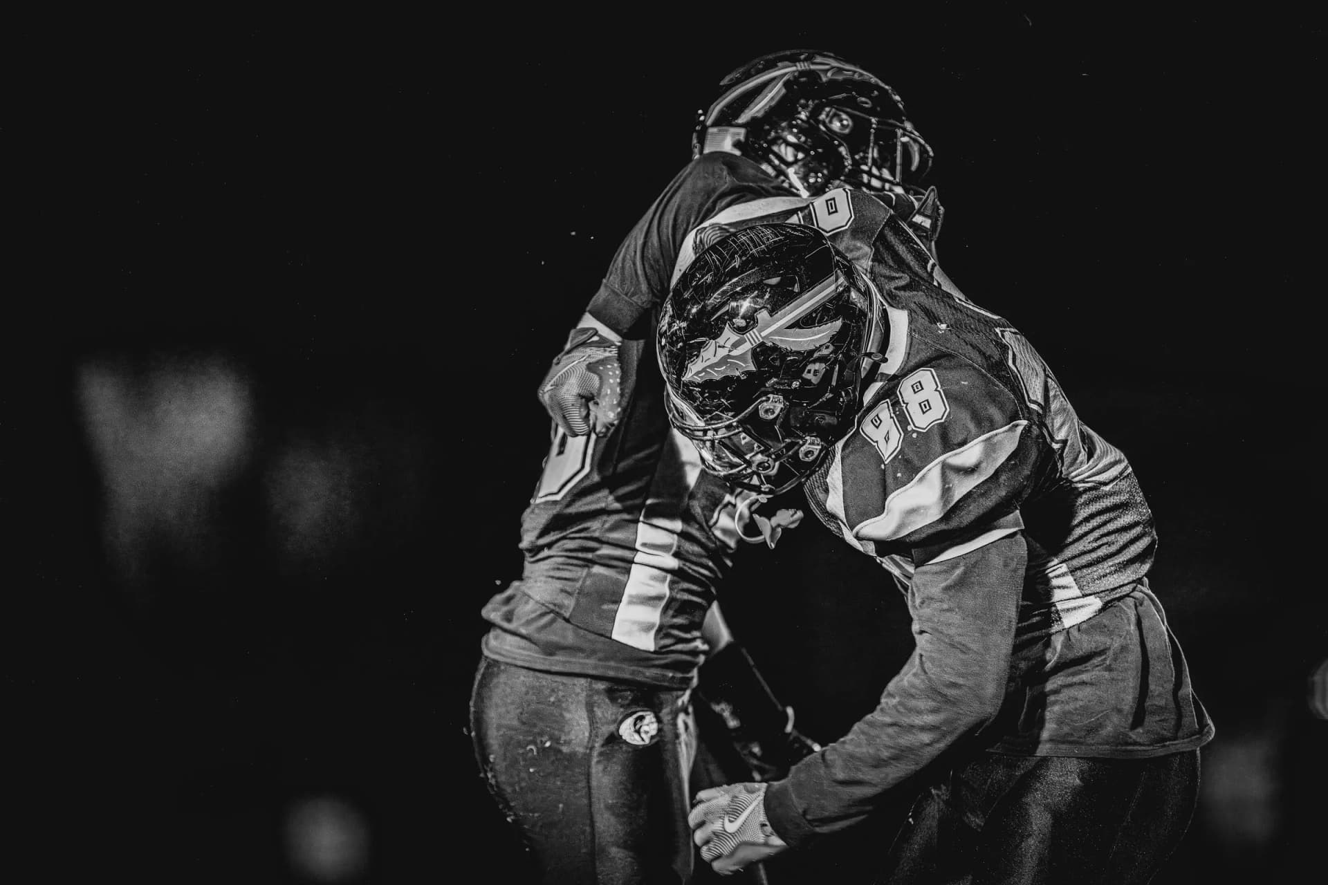 American football players in full gear hug on an outdoor night field, helmets and jerseys lit by artificial lights.
