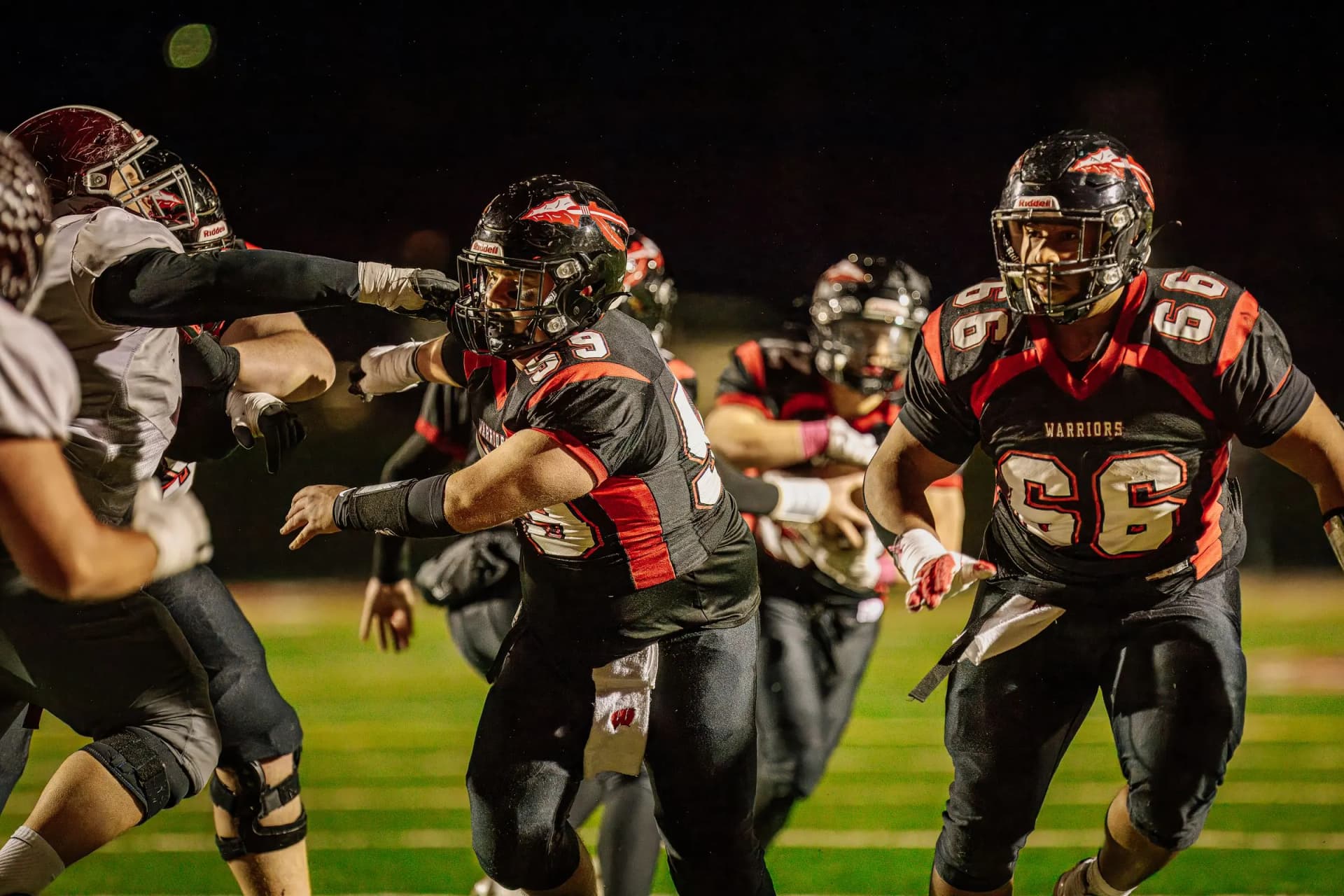 American football players sprint across an outdoor night field illuminated under bright stadium lights.