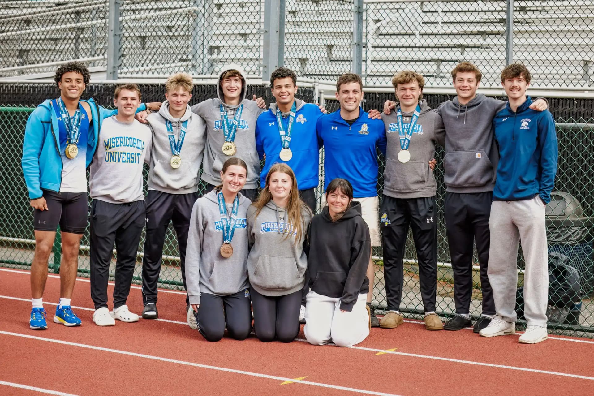 A group of eleven young athletes pose on a red track beside a chain‑link fence and empty bleachers outdoors.