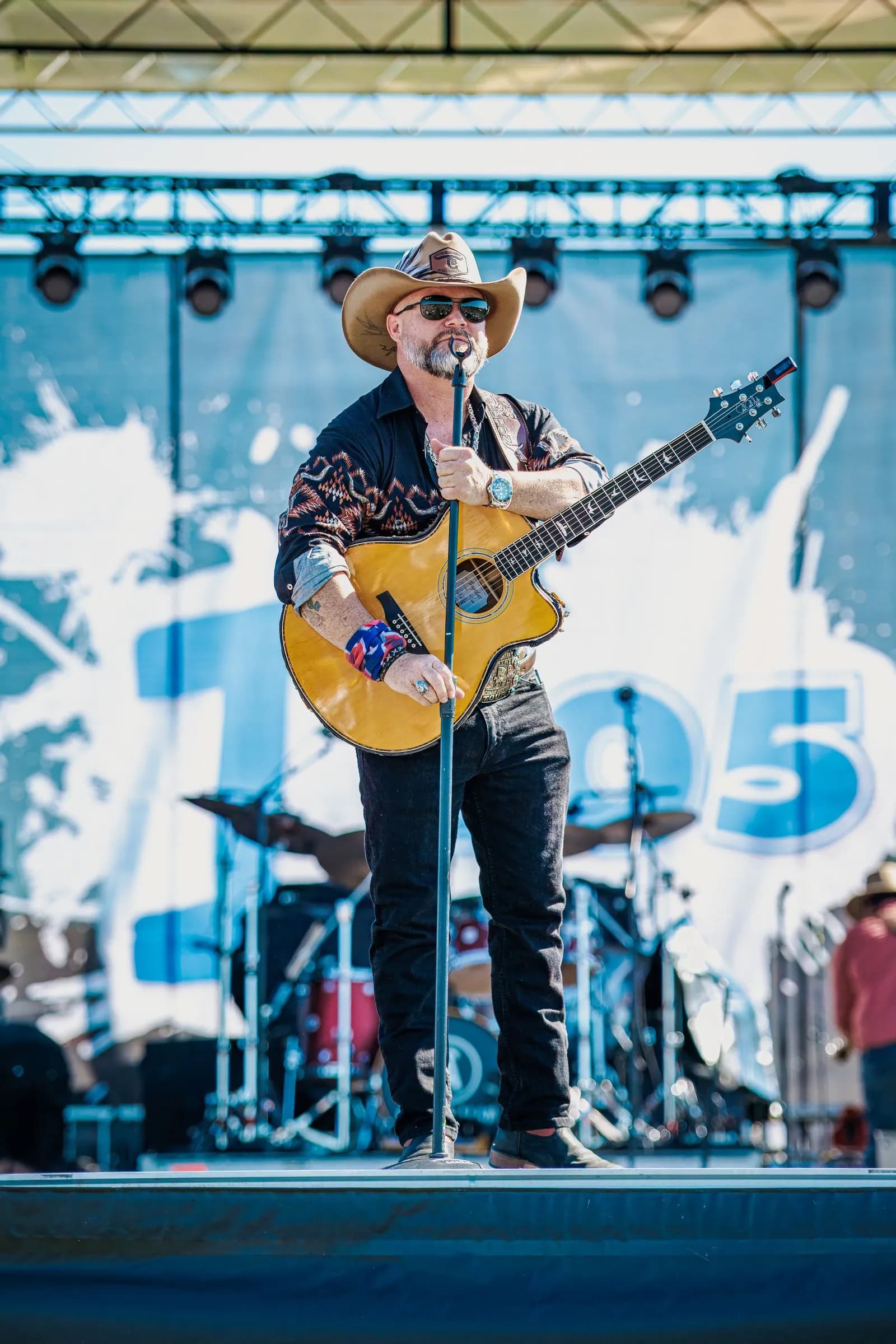 Man in a cowboy hat and sunglasses stands on an outdoor stage at Overlook Park, holding a guitar and a microphone stand.