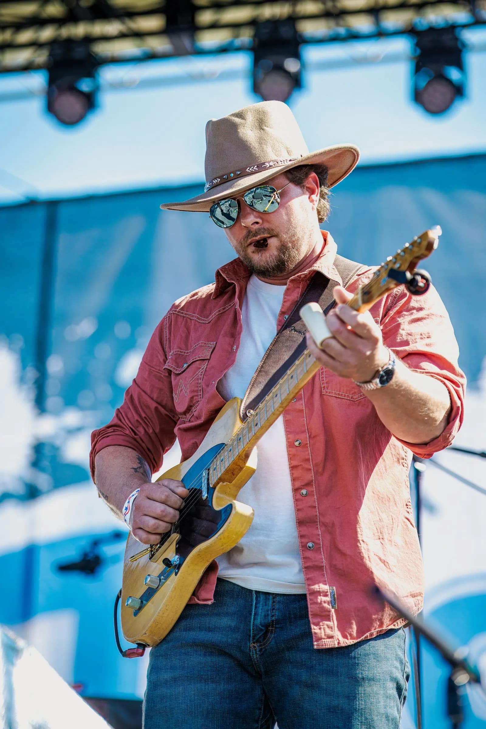 A bearded man in a cowboy hat plays electric guitar on stage at Overlook Park under bright sunlight.