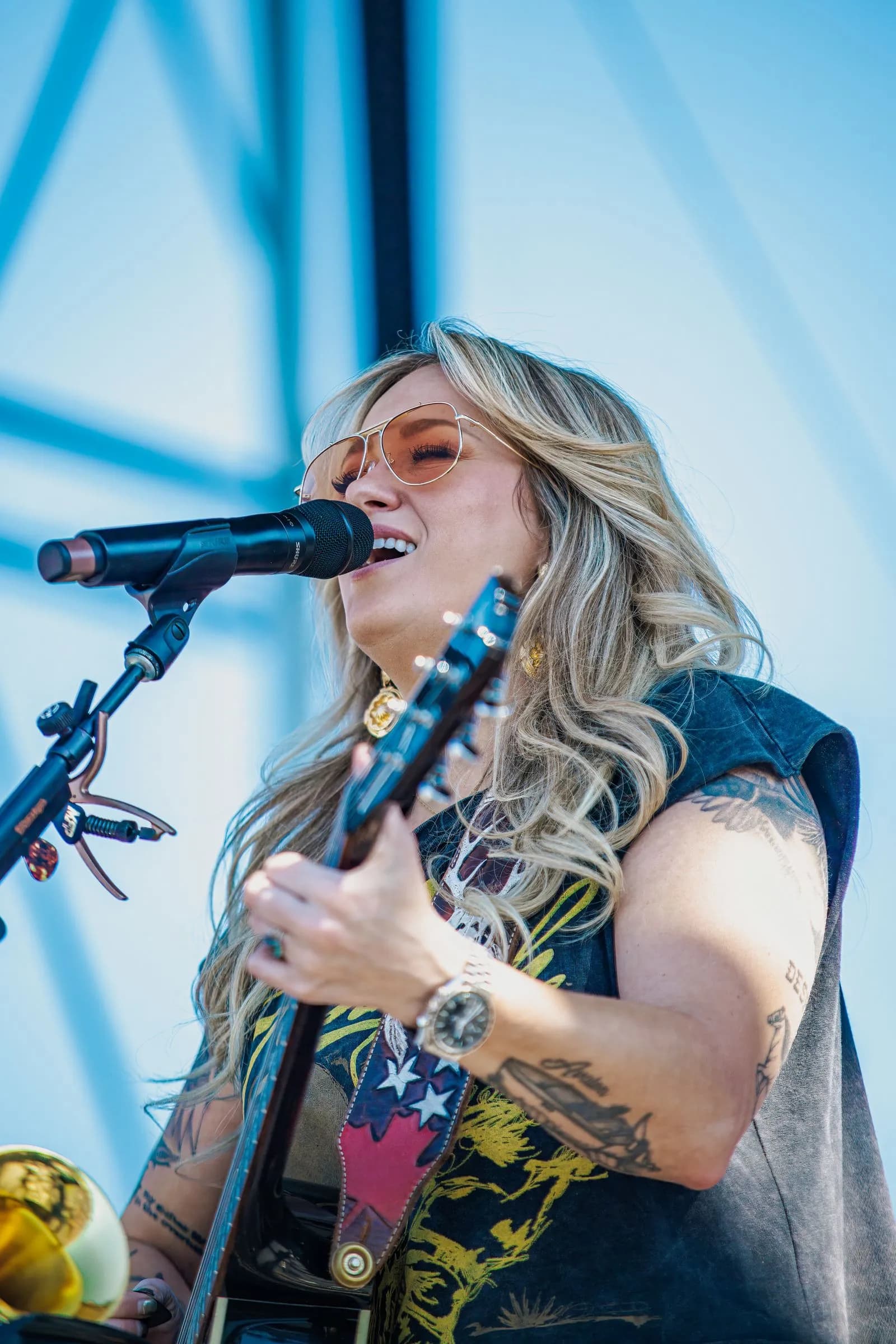 A singer in sunglasses sings into a microphone while holding a guitar on an outdoor stage against a light blue backdrop.