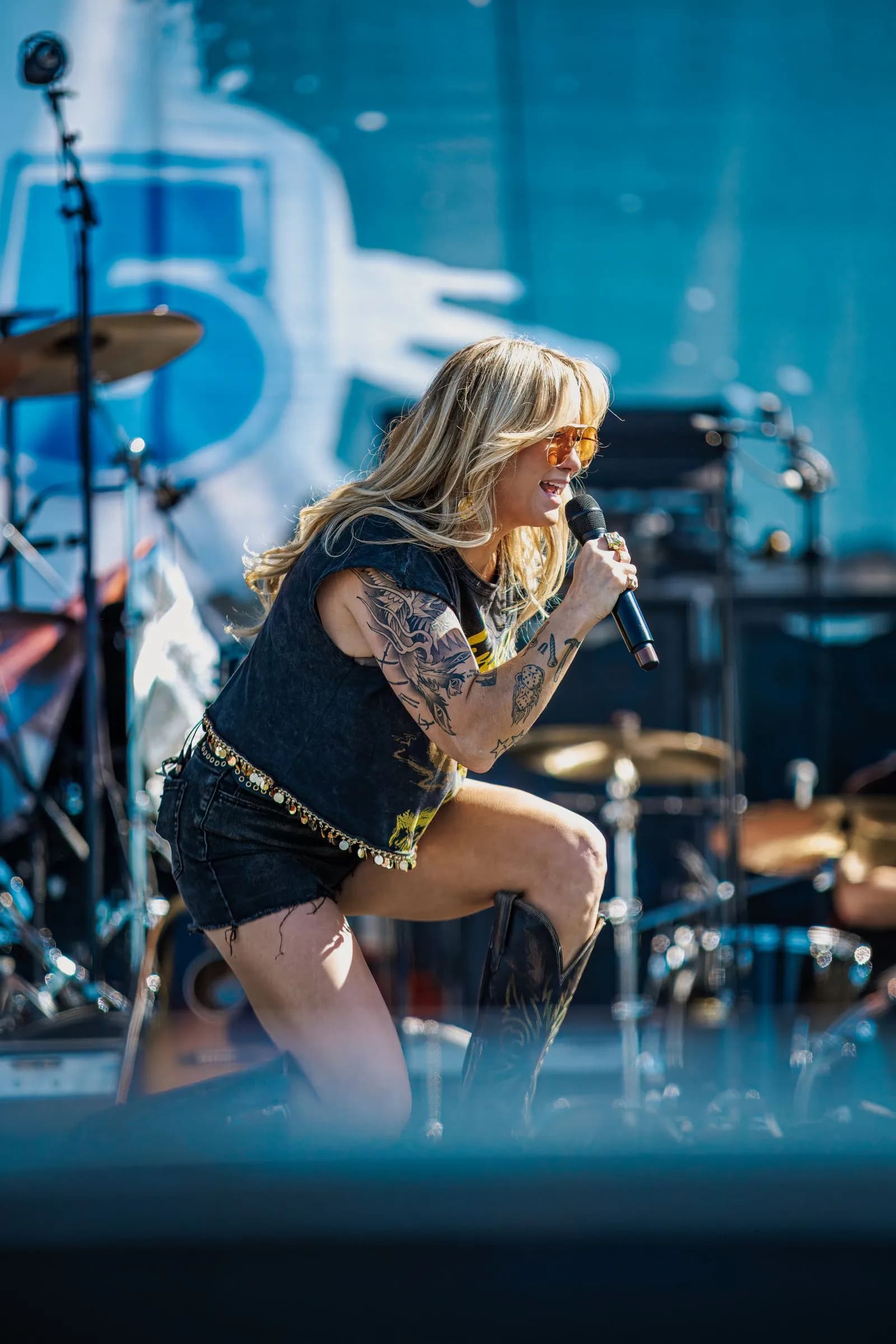 A singer wearing sunglasses and a studded belt kneels on one knee, singing into a microphone outdoors at Overlook Park.
