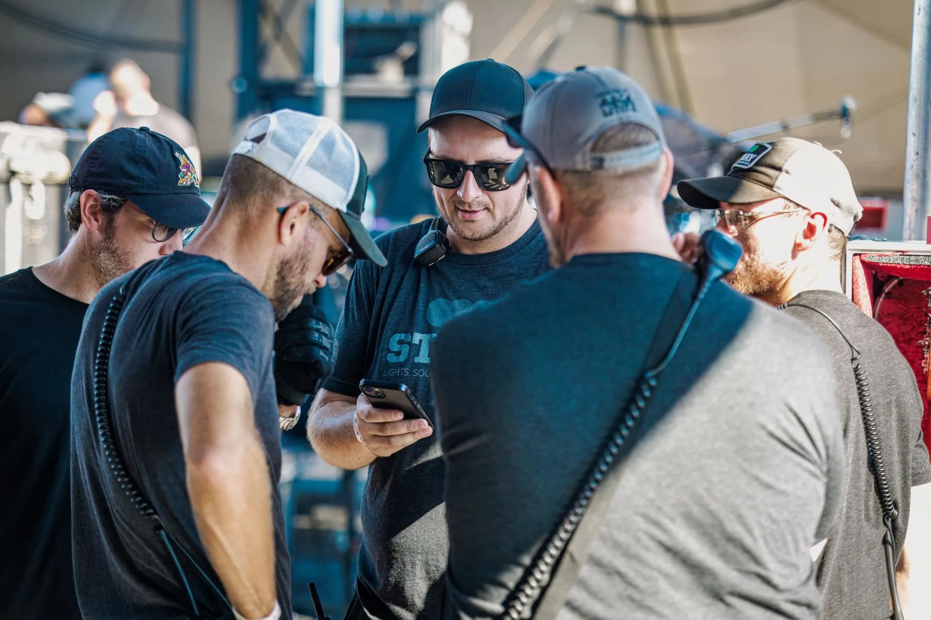 A group of men huddle around a smartphone under bright outdoor light at Overlook Park.