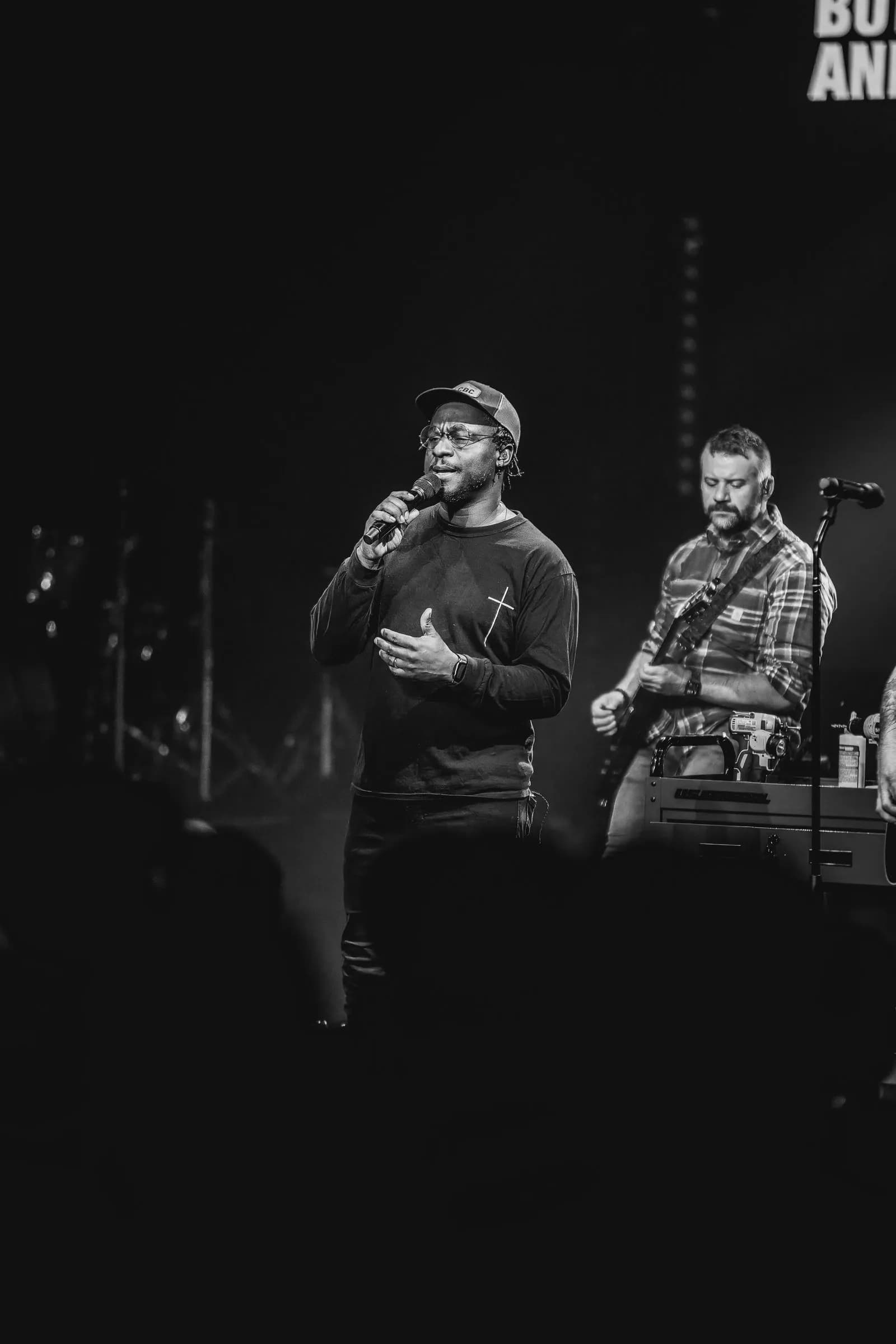 Two men perform on an indoor stage, one singing into a microphone while the other plays electric guitar.