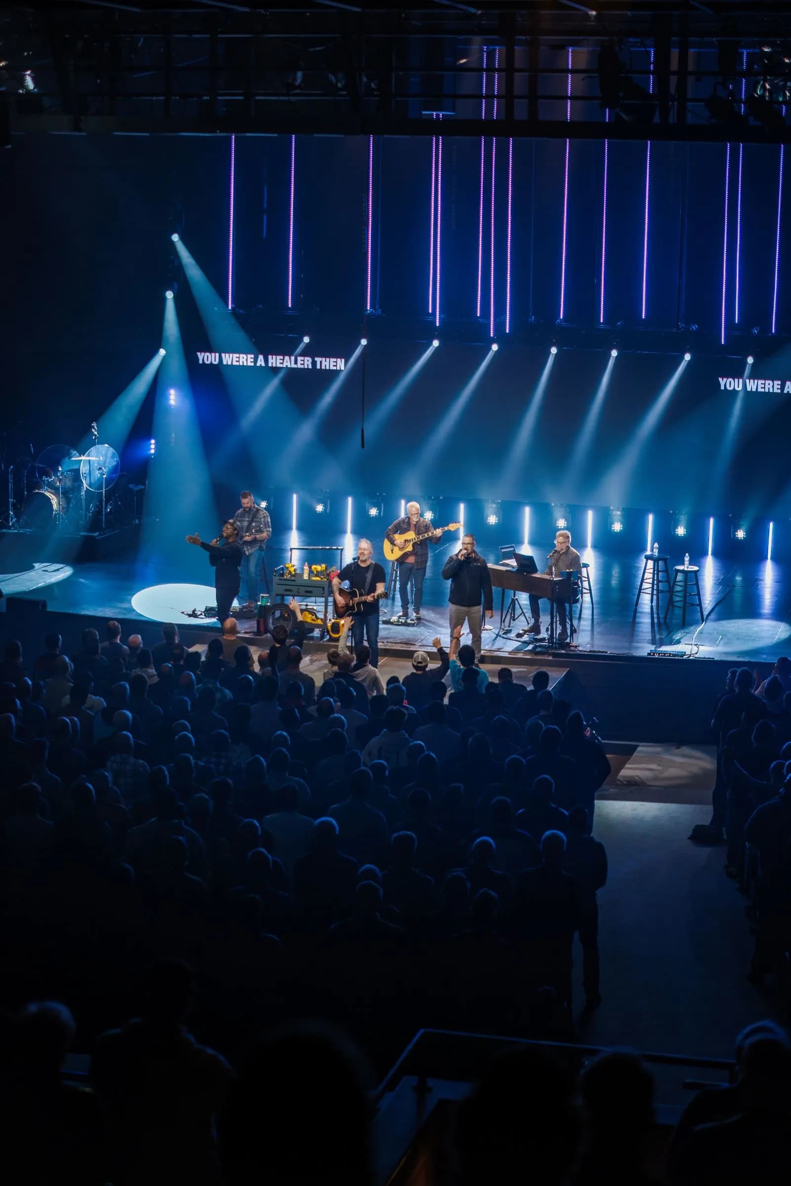 A five‑member band performs on an indoor stage lit by white spotlights and purple LED backdrops.