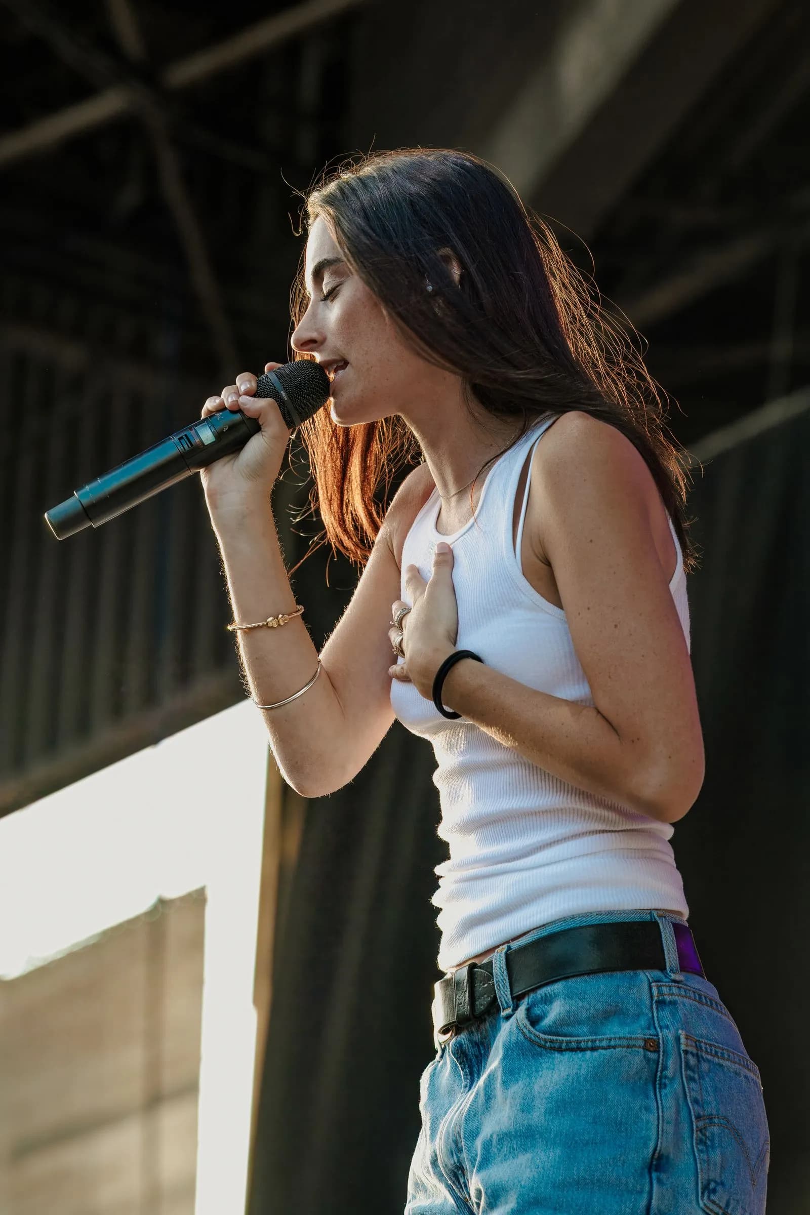 A woman in a white tank top and blue jeans sings into a microphone outdoors at the Summer of Boots Festival